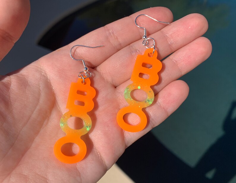 May include: A pair of orange "BOO" earrings with silver hooks. The earrings have a gradient effect, transitioning from orange to yellow-green with glitter accents. The earrings are designed for Halloween.