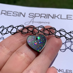 May include: A black choker necklace with a heart-shaped pendant. The pendant is made of resin and has a glittery finish with iridescent colors.