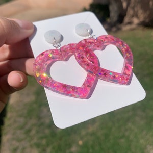 May include: Pink heart-shaped earrings with glitter and white stars. The earrings are on a white card.