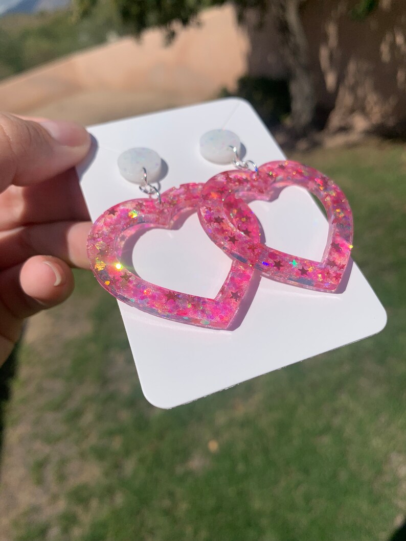 May include: Pink heart-shaped earrings with glitter and white stars. The earrings are on a white card.