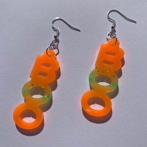 May include: A pair of orange "BOO" earrings with silver hooks. The earrings are made of a translucent material with glitter accents. The bottom half of the earrings have a green and yellow gradient.