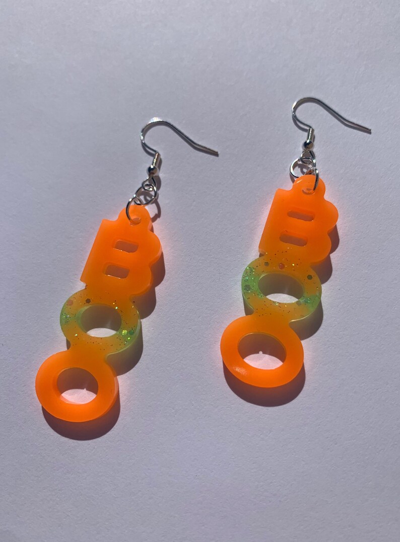 May include: A pair of orange "BOO" earrings with silver hooks. The earrings are made of a translucent material with glitter accents. The bottom half of the earrings have a green and yellow gradient.