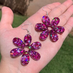 May include: A pair of pink glitter flower earrings. The earrings are made of resin and have a silver hook.
