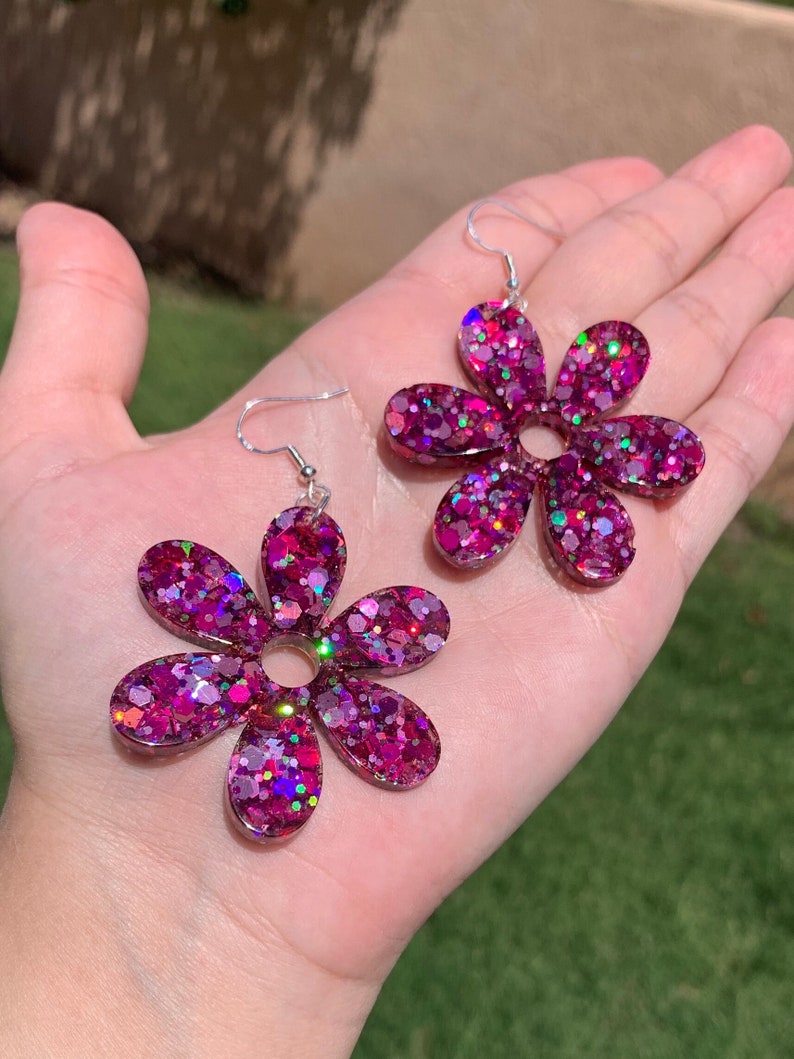 May include: A pair of pink glitter flower earrings. The earrings are made of resin and have a silver hook.