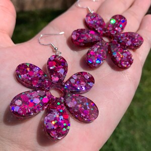 May include: A pair of pink flower-shaped earrings with silver hooks. The earrings are made of resin and have a glittery finish.