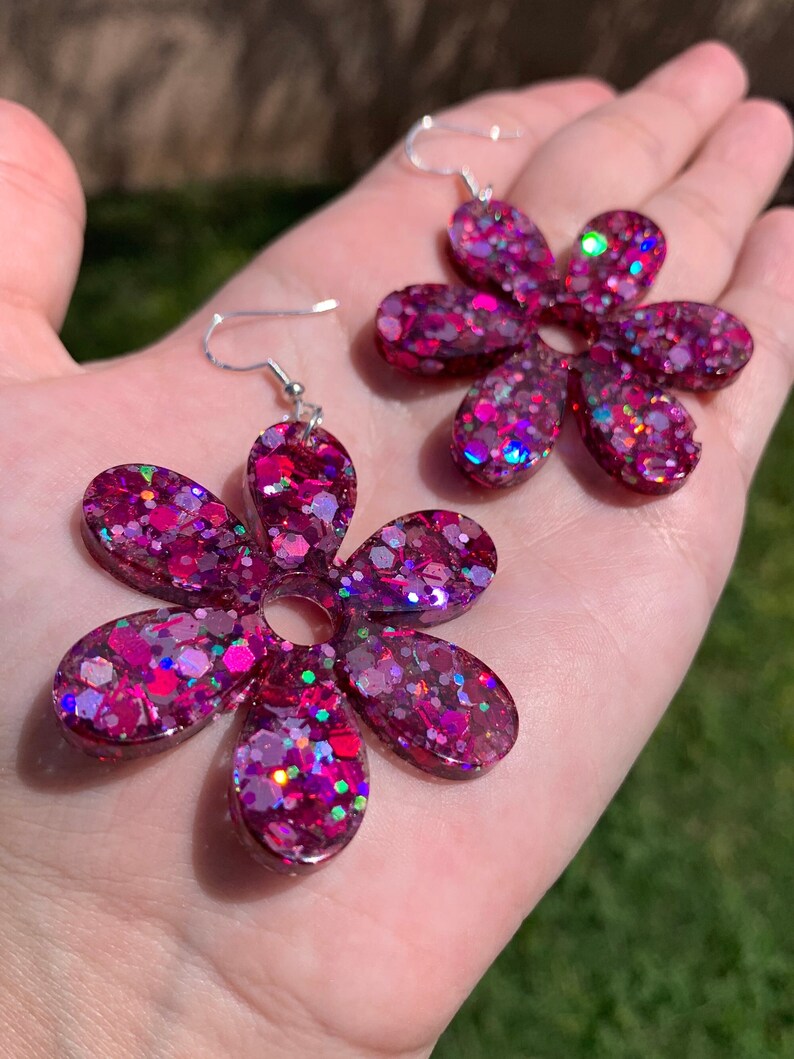May include: A pair of pink flower-shaped earrings with silver hooks. The earrings are made of resin and have a glittery finish.