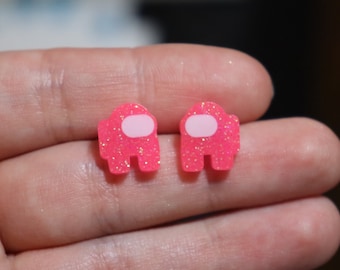 Könnte beinhalten: Ein Paar Ohrringe aus pinkfarbenem Glitzer in Form des Charakters aus dem Videospiel Among Us. Die Ohrringe haben einen weißen Umriss.