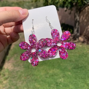 May include: A pair of pink glitter flower dangle earrings with silver hooks. The earrings are made of resin and have a sparkly finish.