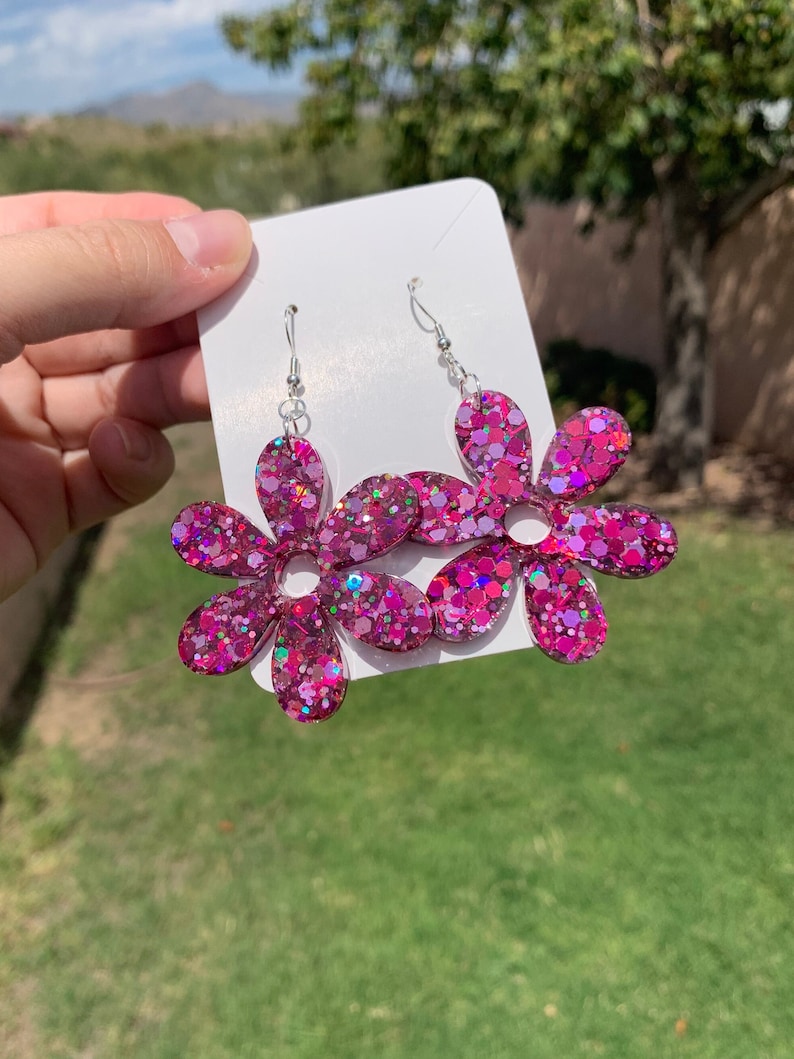 May include: A pair of pink glitter flower dangle earrings with silver hooks. The earrings are made of resin and have a sparkly finish.