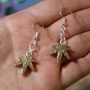 May include: A pair of gold glitter marijuana leaf dangle earrings with silver hooks and chains.