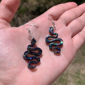 May include: A pair of black snake-shaped earrings with iridescent glitter embedded in the resin. The earrings have silver hooks.