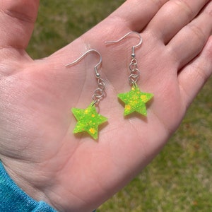 May include: A pair of star-shaped earrings. The earrings are a bright neon green color with yellow glitter accents. They hang from silver-colored fish hook ear wires and small chain links. The earrings are held in an open hand.