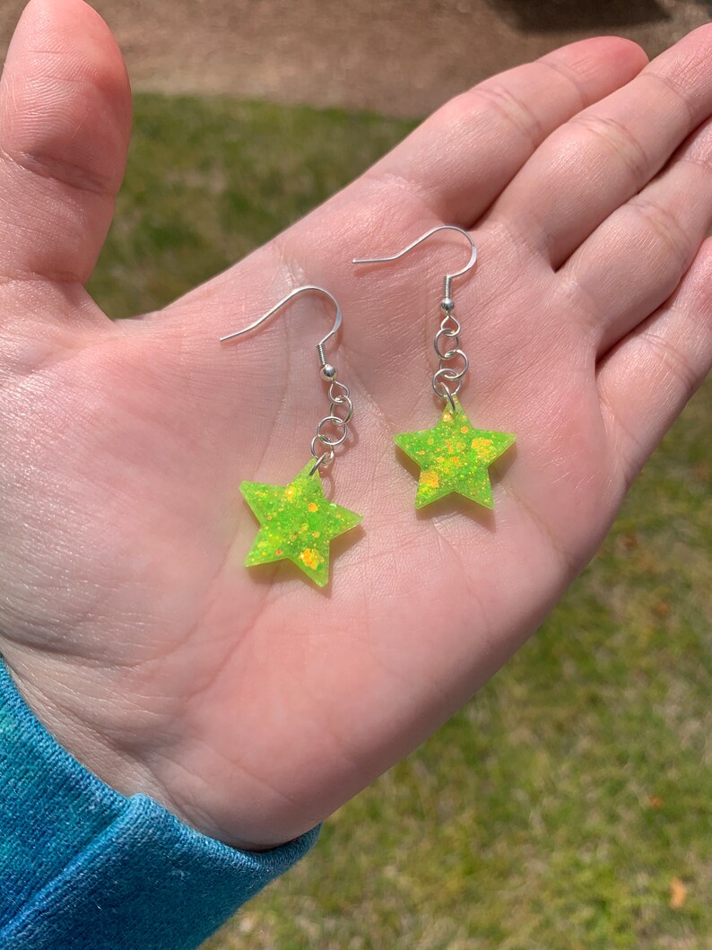May include: A pair of star-shaped earrings. The earrings are a bright neon green color with yellow glitter accents. They hang from silver-colored fish hook ear wires and small chain links. The earrings are held in an open hand.