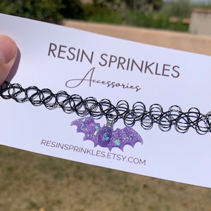 May include: A black choker necklace with a purple glitter bat charm. The charm is made of resin and has a sparkly finish. The text "RESIN SPRINKLES Accessories" and "RESINSPRINKLES.ETSY.COM" is printed on the white card behind the necklace.