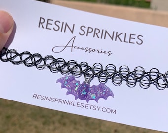 May include: A black choker necklace with a purple glitter bat charm. The charm is made of resin and has a sparkly finish. The text "RESIN SPRINKLES Accessories" and "RESINSPRINKLES.ETSY.COM" is printed on the white card behind the necklace.