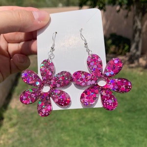 May include: A pair of pink glitter flower dangle earrings. The earrings are made of resin and have a silver hook.