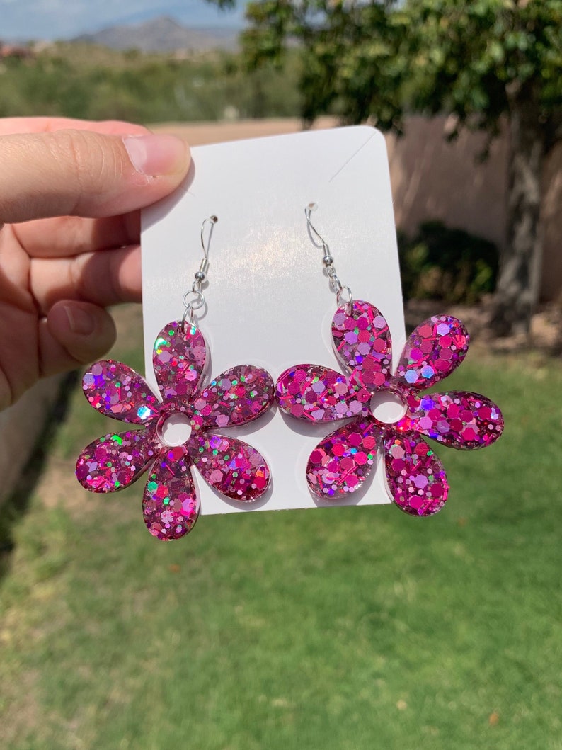 May include: A pair of pink glitter flower dangle earrings. The earrings are made of resin and have a silver hook.