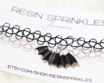 May include: Black choker necklace with a black bat charm. The choker is made of elastic and has a springy design. The bat charm is made of resin and has a detailed design. The text "RESIN SPRINKLES" and "ETSY.COM/SHOP/RESINSPRINKLES" is printed on a white card behind the necklace.