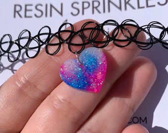May include: A black choker necklace with a pink, blue, and white glitter heart charm. The charm is made of resin and has a sparkly finish.
