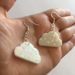 May include: A pair of white cloud-shaped earrings with iridescent glitter. The earrings are made of resin and have gold hooks.