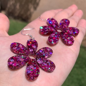 May include: A pair of pink glitter flower earrings with silver hooks. The flowers are made of resin and have a sparkly finish.