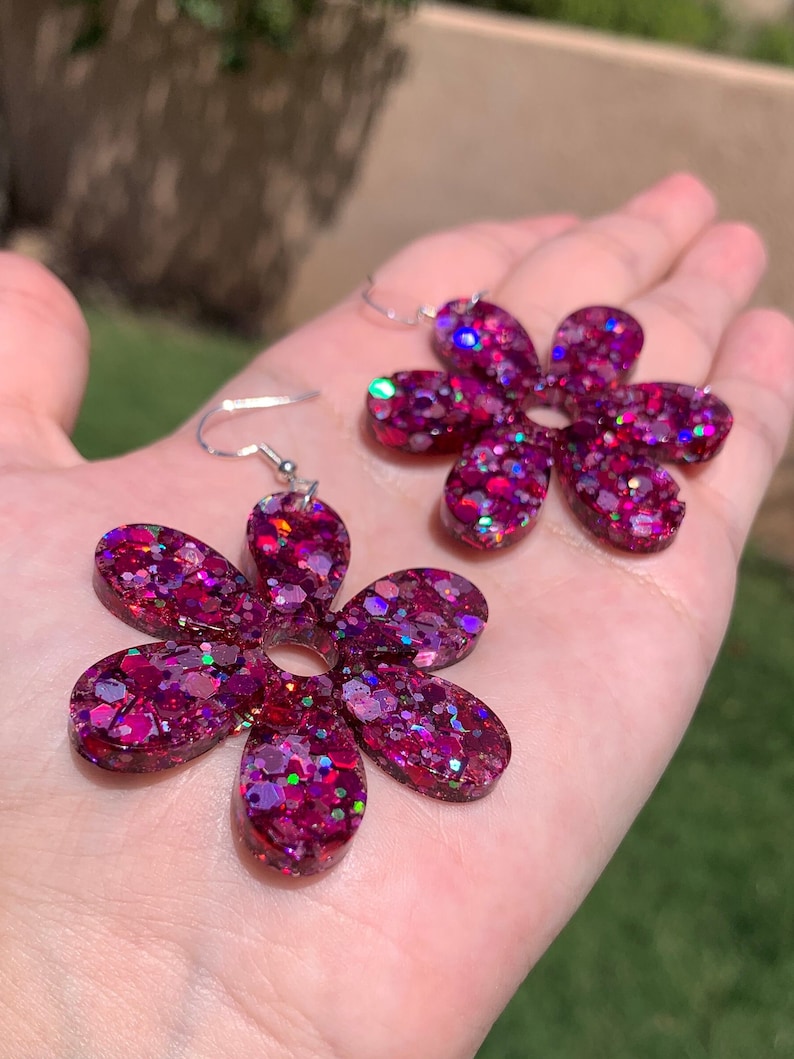 May include: A pair of pink glitter flower earrings with silver hooks. The flowers are made of resin and have a sparkly finish.