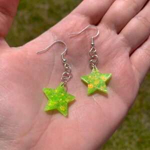 May include: A pair of star-shaped earrings. Each earring features a neon green star with glitter accents, suspended from a silver-colored hook and chain. The earrings are held in a hand against a blurred green background.