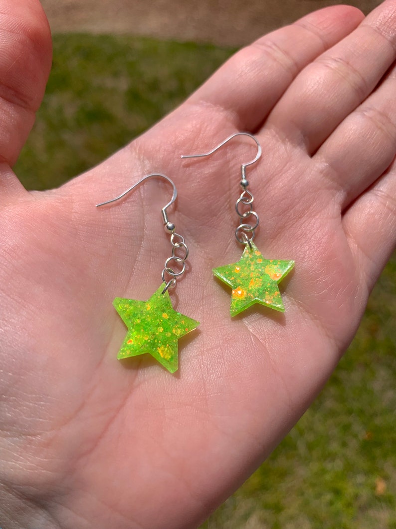 May include: A pair of star-shaped earrings. Each earring features a neon green star with glitter accents, suspended from a silver-colored hook and chain. The earrings are held in a hand against a blurred green background.