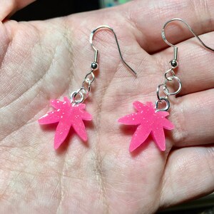 May include: A pair of silver dangle earrings with pink glitter leaf charms. The leaves are a stylized marijuana leaf shape.