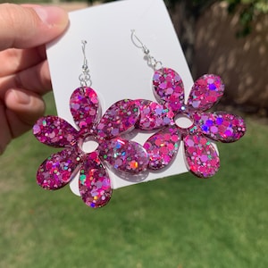 May include: A pair of pink glitter flower earrings with silver hooks. The flowers are made of resin and have a sparkly finish.