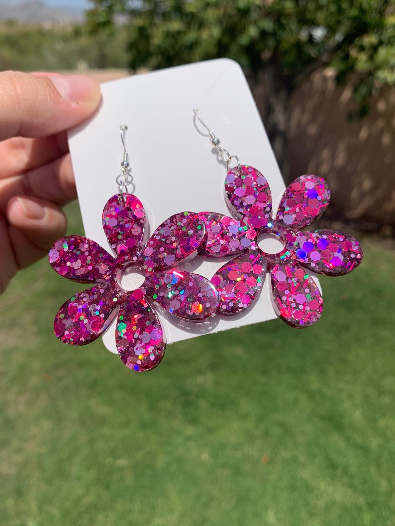 May include: A pair of pink glitter flower earrings with silver hooks. The flowers are made of resin and have a sparkly finish.