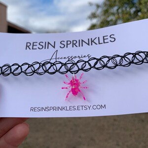 May include: A black choker necklace with a pink glitter spider charm. The charm is made of resin and has a sparkly finish. The necklace is displayed on a white card with the text "RESIN SPRINKLES Accessories" and "RESINSPRINKLES.ETSY.COM".