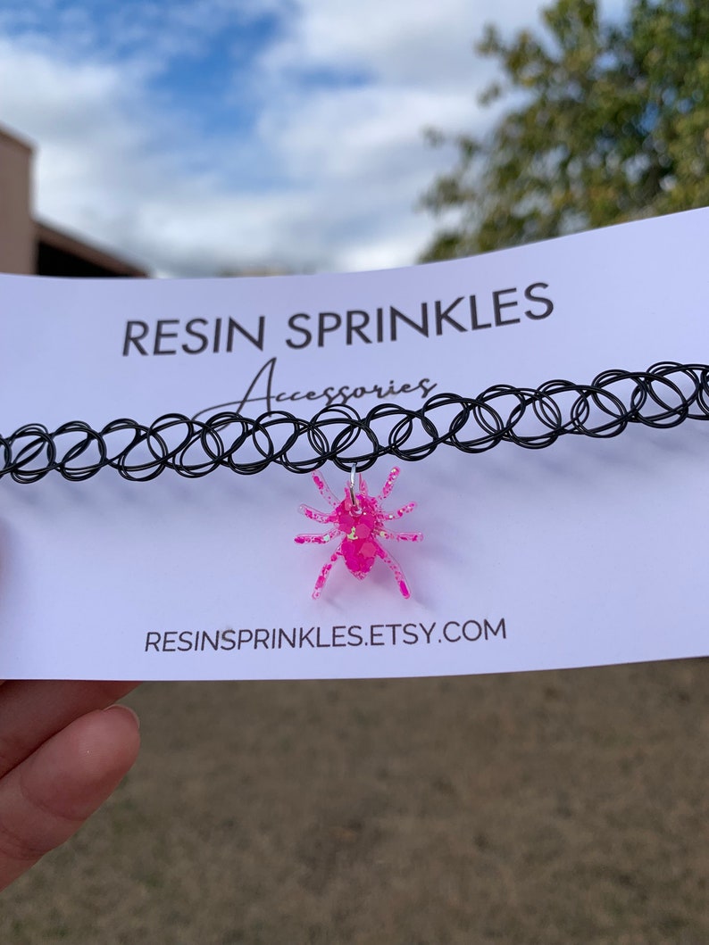 May include: A black choker necklace with a pink glitter spider charm. The charm is made of resin and has a sparkly finish. The necklace is displayed on a white card with the text "RESIN SPRINKLES Accessories" and "RESINSPRINKLES.ETSY.COM".