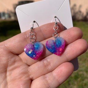 May include: A pair of heart-shaped earrings with a pink, blue, and white glitter design. The earrings are made of resin and have silver hooks.