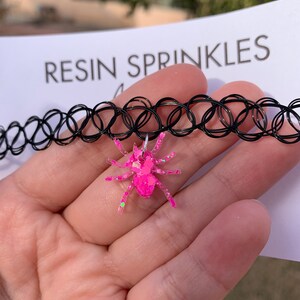 May include: A pink glitter spider charm attached to a black choker necklace. The necklace is made of elastic and has a series of small, connected circles. The charm is made of resin and has a sparkly finish.