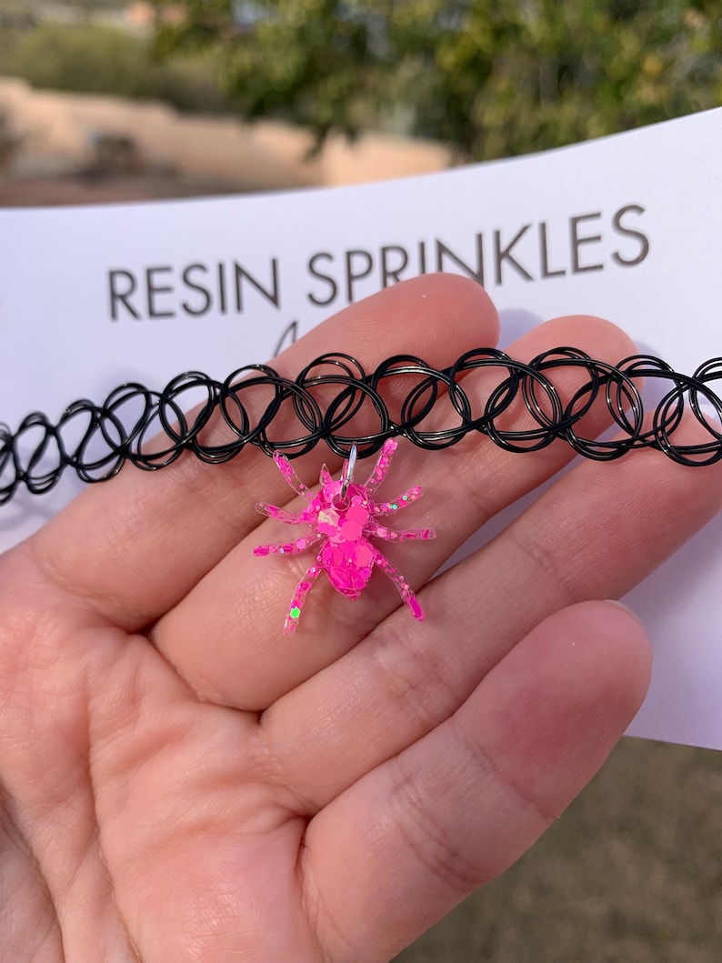 May include: A pink glitter spider charm attached to a black choker necklace. The necklace is made of elastic and has a series of small, connected circles. The charm is made of resin and has a sparkly finish.