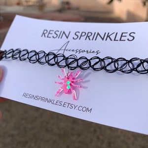 May include: A black choker necklace with a pink resin spider charm. The spider has green glitter accents and is attached to the choker with a silver chain. The text "RESINSPRINKLES.ETSY.COM" is printed on the white card behind the necklace.