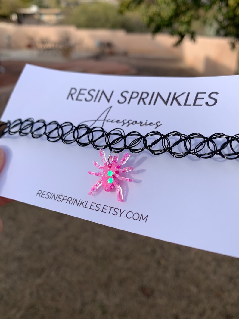 May include: A black choker necklace with a pink resin spider charm. The spider has green glitter accents and is attached to the choker with a silver chain. The text "RESINSPRINKLES.ETSY.COM" is printed on the white card behind the necklace.