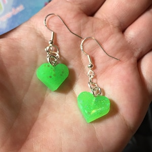 May include: A pair of silver heart-shaped earrings with green glitter. The hearts are small and dangle from silver hooks.