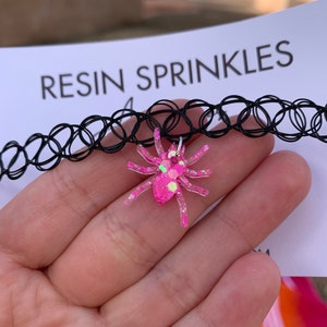 May include: A black choker necklace with a pink glitter spider charm. The charm is made of resin and has a sparkly finish. The text "RESIN SPRINKLES" is visible in the background.