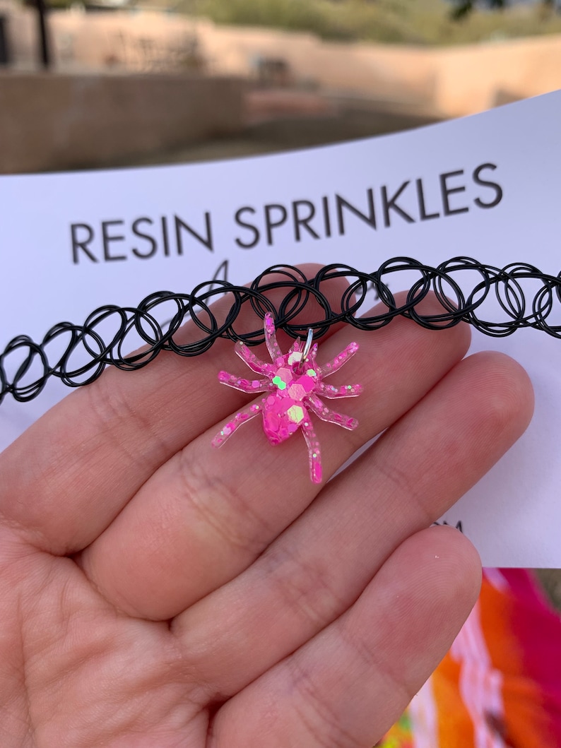 May include: A black choker necklace with a pink glitter spider charm. The charm is made of resin and has a sparkly finish. The text "RESIN SPRINKLES" is visible in the background.