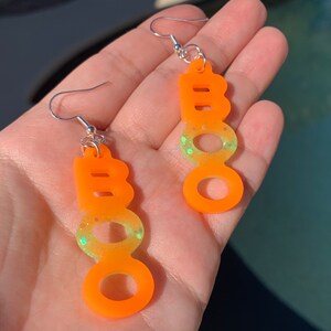 May include: A pair of orange "BOO" earrings with silver hooks. The earrings have a gradient effect, transitioning from orange to a lighter green with glitter. The earrings are designed for Halloween.