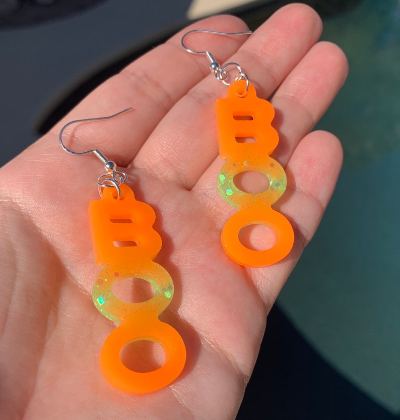 May include: A pair of orange "BOO" earrings with silver hooks. The earrings have a gradient effect, transitioning from orange to a lighter green with glitter. The earrings are designed for Halloween.