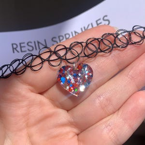 Könnte beinhalten: Schwarze Tattoo-Choker-Halskette mit herzförmigem Anhänger. Der Anhänger ist mit roten, weißen und blauen Glitzer gefüllt. Der Text "RESIN SPRINKLES" ist im Hintergrund zu sehen.