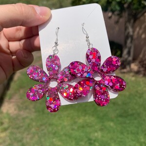 May include: A pair of pink glitter flower earrings with silver hooks. The flowers have a 7-pointed star shape with a hole in the centre.
