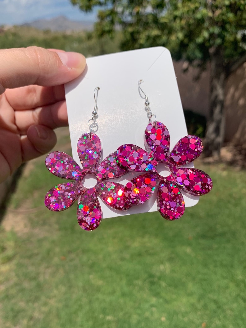 May include: A pair of pink glitter flower earrings with silver hooks. The flowers have a 7-pointed star shape with a hole in the centre.