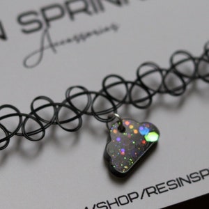May include: A black choker necklace with a silver charm in the shape of a cloud. The cloud charm is filled with iridescent glitter and has a rainbow effect.