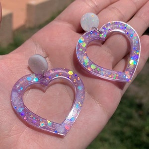 May include: A pair of purple heart-shaped earrings with iridescent glitter embedded in the resin. The earrings have a white circle at the top.