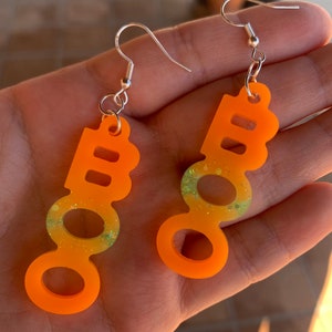 May include: A pair of orange acrylic earrings with a 'BOO' design. The earrings have a gradient of yellow and green glitter in the center. Each earring has a silver-colored hook.