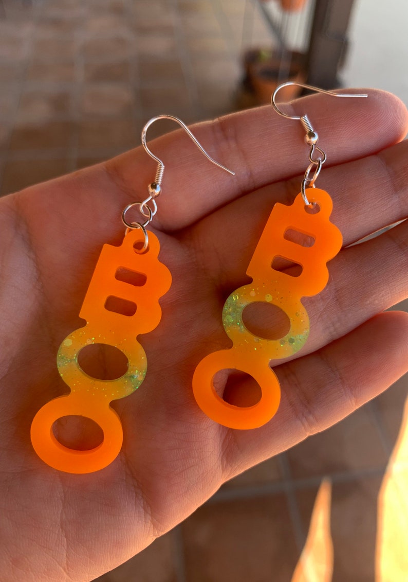 May include: A pair of orange acrylic earrings with a 'BOO' design. The earrings have a gradient of yellow and green glitter in the center. Each earring has a silver-colored hook.